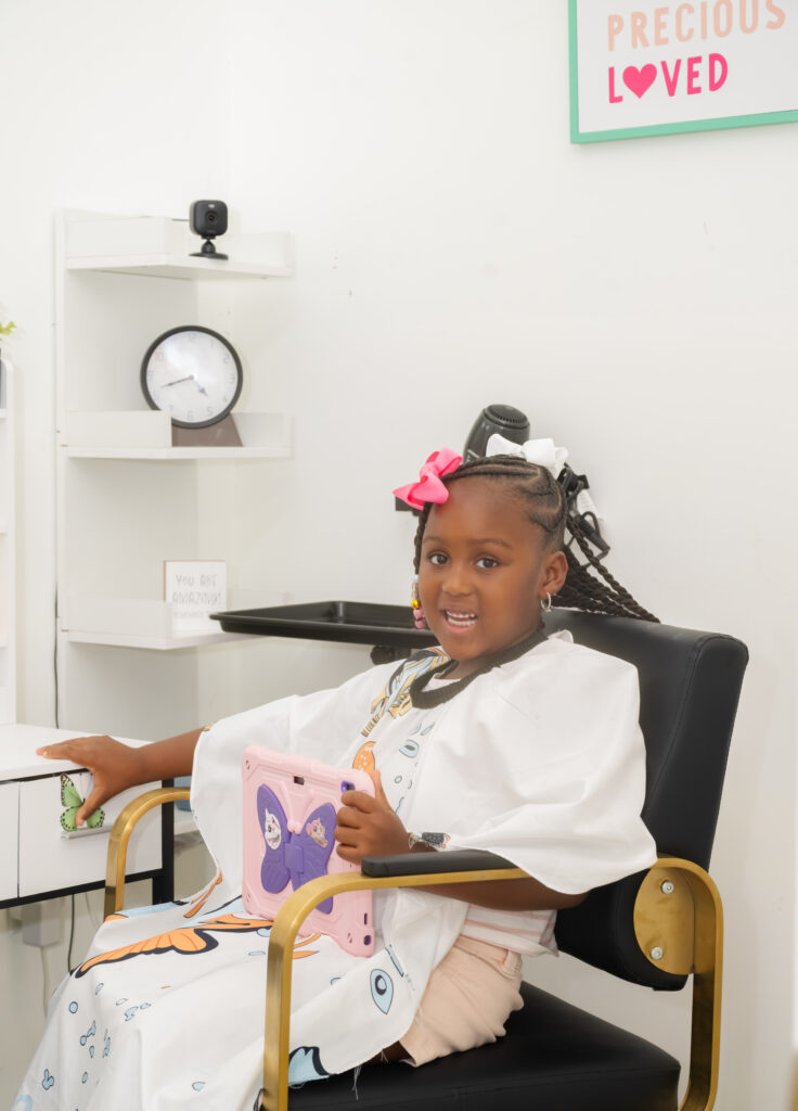 Young child in a styling chair with braided pigtails finished with pink bows, photographed at Nerielle Braiding Studio